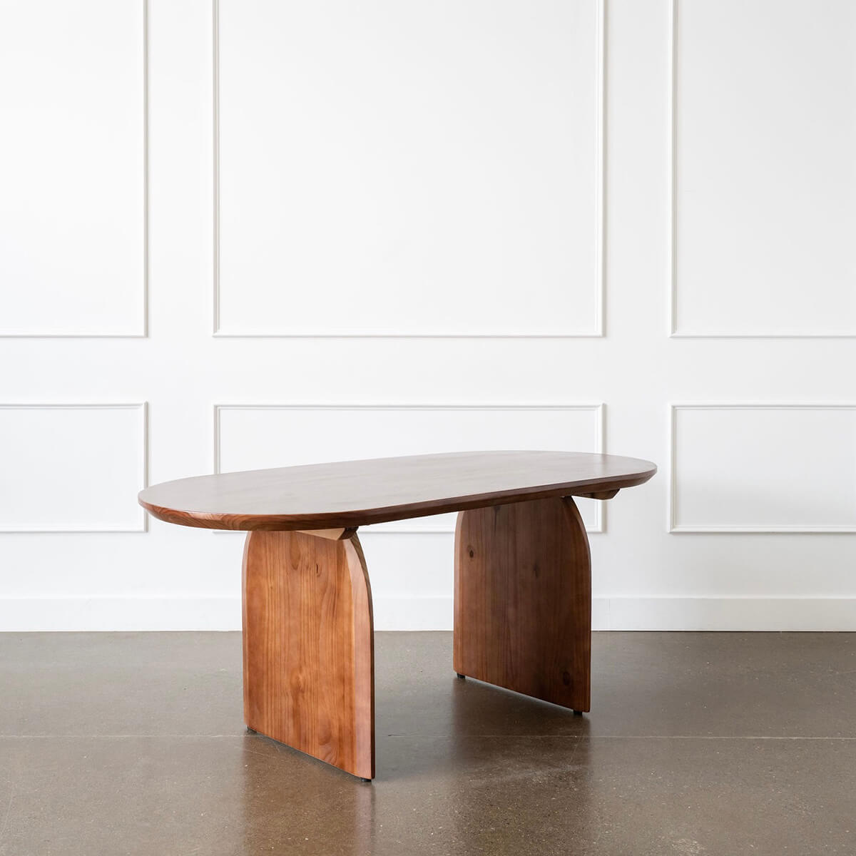 An oval-shaped, walnut dining table with sculptural half-oval slab legs, accompanied by four chairs with black seats and wooden legs. The table has a smooth, honey-toned finish and is set in a room with a white wall and a large window.
